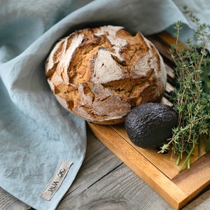 May include: A loaf of sourdough bread on a wooden chopping board with a sprig of thyme and an avocado. The bread is on a blue linen cloth with a tag that says "LAUA_TTC".