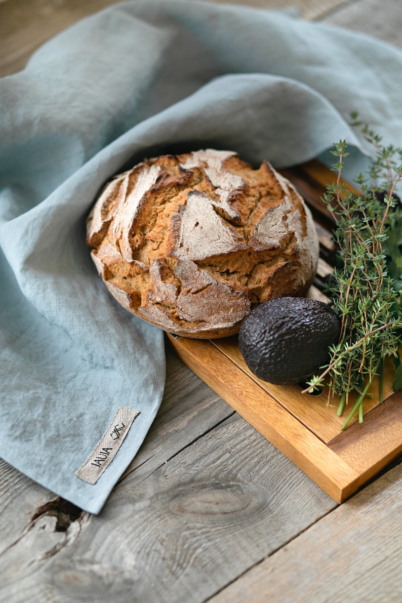 May include: A loaf of sourdough bread on a wooden chopping board with a sprig of thyme and an avocado. The bread is on a blue linen cloth with a tag that says "LAUA_TTC".