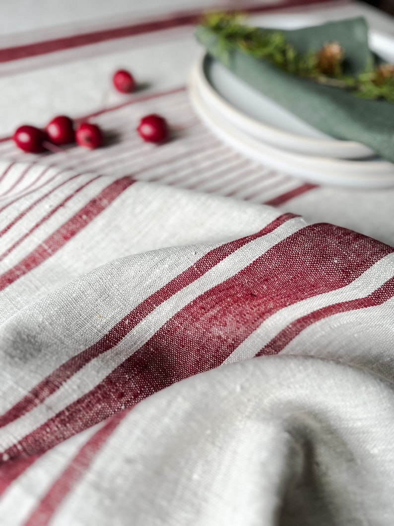 K&ouml;nnte beinhalten: Eine wei&szlig;e Tischdecke aus Leinen mit roten Streifen. Die Tischdecke ist in der Mitte gefaltet und hat ein paar rote Beeren darauf verstreut.