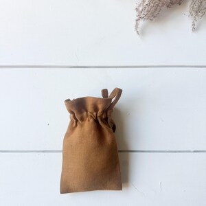 Puede incluir: Una peque&ntilde;a bolsa marr&oacute;n con cord&oacute;n hecha de tela texturizada. La bolsa est&aacute; fruncida en la parte superior y tiene un lazo a cada lado. El fondo es una superficie de madera blanca con una ramita de plantas secas.