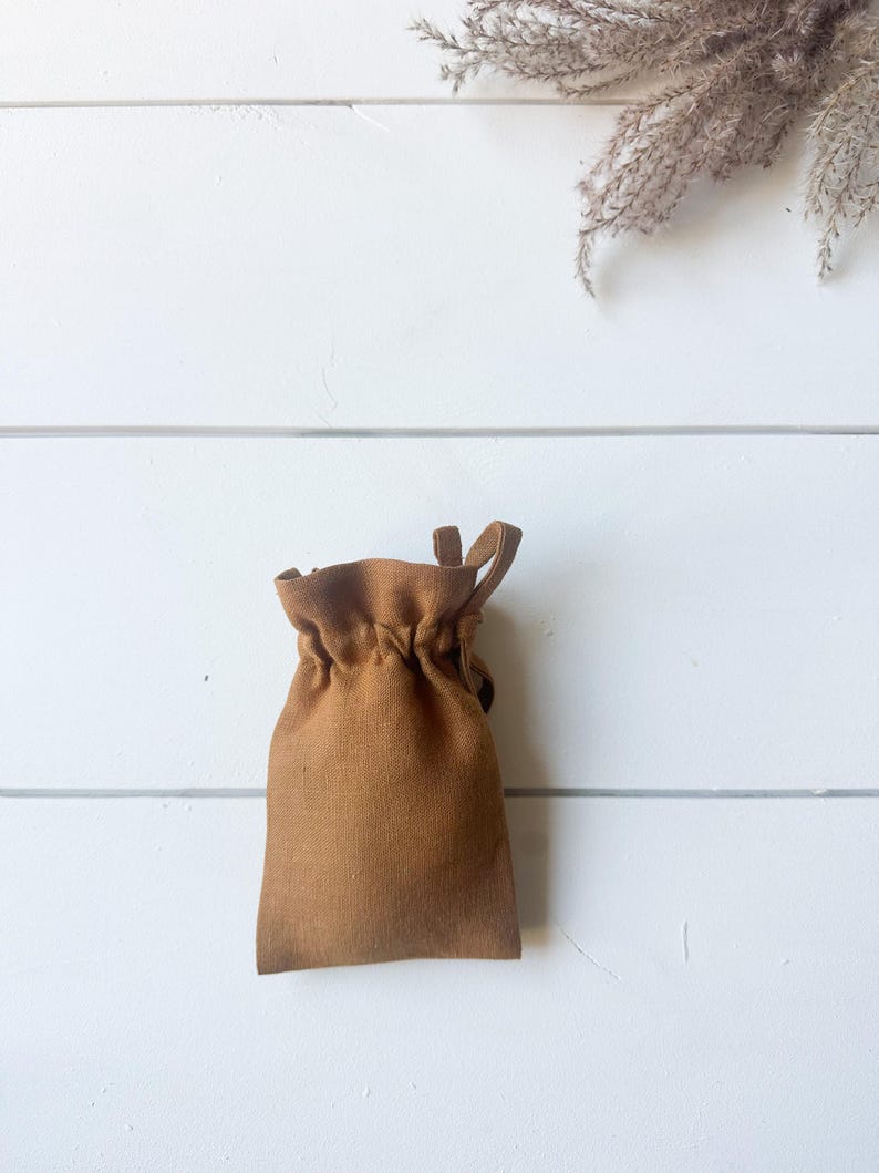 Puede incluir: Una peque&ntilde;a bolsa marr&oacute;n con cord&oacute;n hecha de tela texturizada. La bolsa est&aacute; fruncida en la parte superior y tiene un lazo a cada lado. El fondo es una superficie de madera blanca con una ramita de plantas secas.