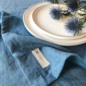 May include: A blue linen tablecloth with a white plate and a bouquet of blue thistle flowers. The tablecloth has a small fabric tag with the symbol "X" on it.