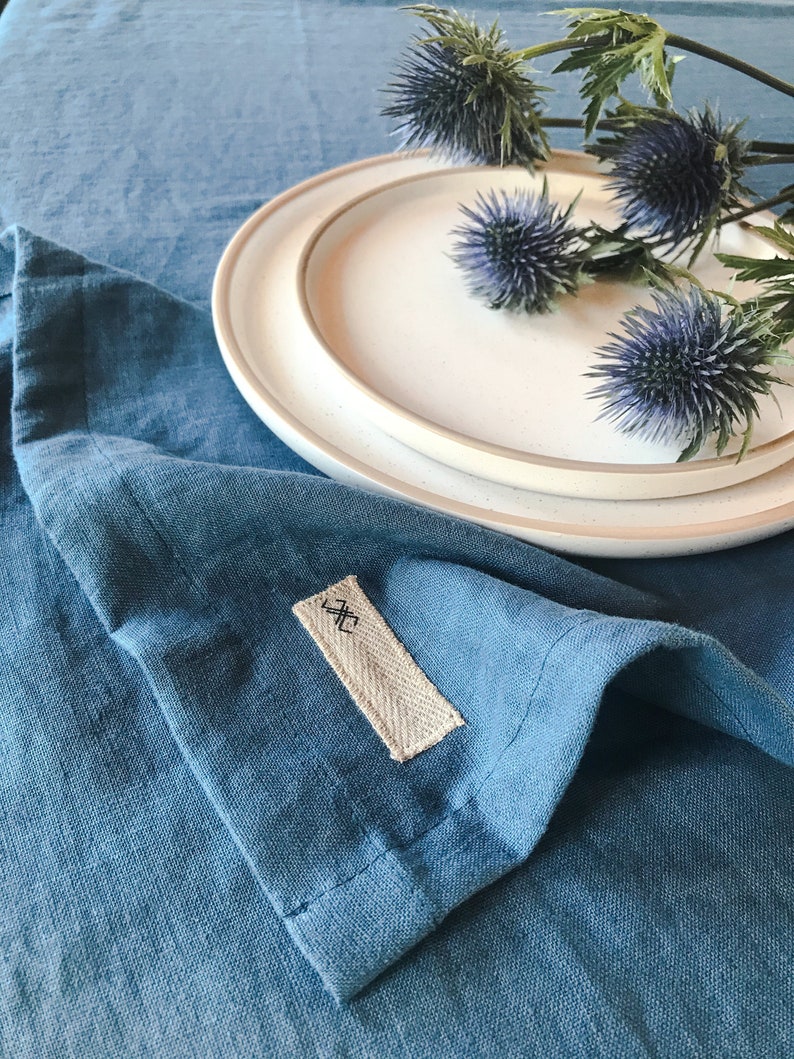 May include: A blue linen tablecloth with a white plate and a bouquet of blue thistle flowers. The tablecloth has a small fabric tag with the symbol "X" on it.