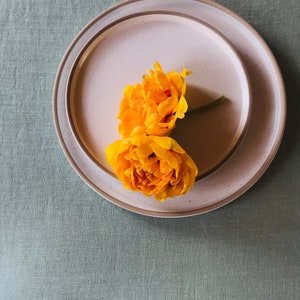 Puede incluir: Dos flores amarillas sobre un plato de cer&aacute;mica blanca con borde marr&oacute;n. El plato est&aacute; sobre un mantel de lino verde.