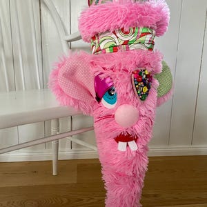 Könnte beinhalten: Ein skurriler rosa Plüsch-Hasen-Strumpf mit flauschiger Textur. Der Hase hat eine rosa Nase, ein blaues Auge und einen Mund mit zwei weißen Zähnen. Der Strumpf ist mit gemustertem Stoff und bunten Verzierungen verziert.