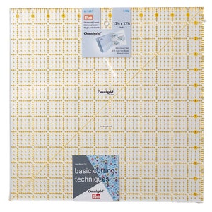 May include: A clear Omnigrid quilting ruler, measuring 31.75 x 31.75 cm, with yellow grid lines and markings. Includes a handbook for basic cutting techniques. The ruler has a diagonal line for angle measurements and is packaged in clear plastic.
