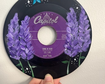 以下が含まれることがあります： 紫の花と青い蝶が描かれた黒いビニールレコード。レコードのラベルは、キャピトルレコードのロゴと、レス・ポールとメアリー・フォードによる「ソング・イン・ブルー」という曲名が書かれた紫色のラベルです。