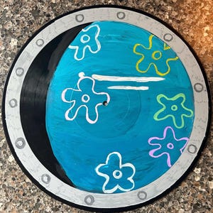May include: A black vinyl record painted to look like a porthole with a blue background and white, yellow, pink, and green flowers.