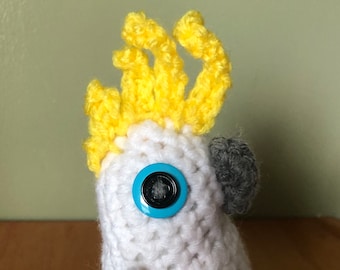 May include: A white crocheted cockatoo plush toy featuring a bright yellow crest, a blue button eye, and a dark gray beak and feet. The bird is crafted with visible yarn stitches and maintains a standing posture with its wings tucked at its sides.