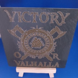 Könnte beinhalten: Eine schwarze Schiefertafel mit goldener Beschriftung, die "VICTORY OVER VALHALLA" lautet. Die Tafel zeigt ein kreisförmiges Design mit einem Symbol eines Dreiecks mit einem Kreis im Inneren, umgeben von Runen. Zwei gekreuzte Äxte befinden sich hinter dem Kreis.