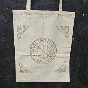 Könnte beinhalten: Eine weiße Stofftasche mit braunen Druckdesigns. Die Tasche hat ein kreisförmiges Design mit Runen und einem Symbol in der Mitte. Die Tasche hat auch vier Eckdesigns.