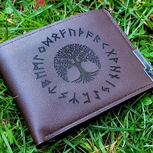 Puede incluir: Una cartera de cuero marrón con un diseño de árbol de la vida y runas nórdicas alrededor del borde. La cartera tiene un acento de metal plateado en el lateral.