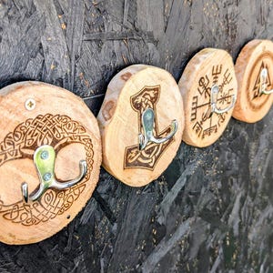 May include: Four wooden wall hooks with different Norse designs, including a tree of life, a hammer, a compass, and a triangle. Each hook has a silver metal double hook.