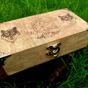 May include: A wooden box with a hinged lid and a brass latch. The box is decorated with a wood-burned design of two wolf heads and a compass.