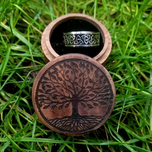 May include: A wooden ring box with a tree of life design on the lid. The box is open and a silver ring with a Celtic knot design is inside.