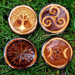 May include: Four wooden coasters with different symbols carved into them. The coasters are round and have a natural wood finish. The symbols include a tree of life, a triskelion, a compass rose, and a triple moon with a pentagram.