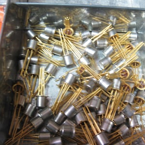 Puede incluir: Primer plano de una gran cantidad de componentes electrónicos de color dorado, probablemente transistores, en un contenedor metálico. Los componentes están dispuestos en un patrón aleatorio y parecen ser nuevos o sin usar.