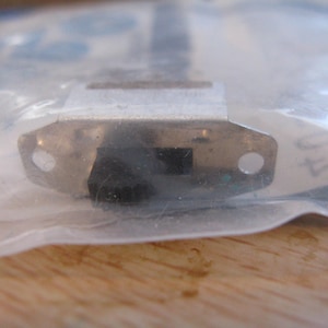 Puede incluir: Un pequeño componente metálico con un botón rectangular negro y dos agujeros circulares. El componente está dentro de una bolsa de plástico transparente, con una superficie de madera visible en el fondo.