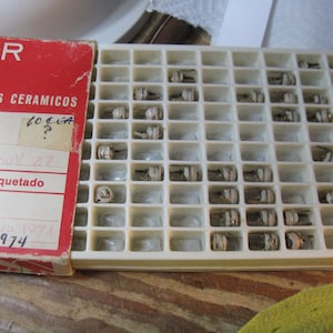 Puede incluir: Una bandeja de plástico blanco con filas de pequeños condensadores cerámicos. La bandeja está etiquetada con "Condensadores Ceramicos" y "Empaquetado". La fecha "1974" está escrita en la etiqueta.