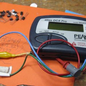 May include: A blue and black electronic device with a screen that reads "atlas DCA Pro" and "DCA75". The device has a red, blue, and green wire attached to it. The device is on a brown surface with a piece of paper that has writing on it.