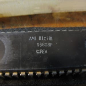 May include: Close-up of a black integrated circuit chip with the text "AMI 8107BL S6808P KOREA" printed on the surface. The chip has numerous gold-colored pins along the bottom edge.