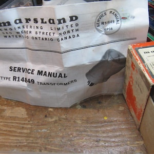 Peut inclure: Un manuel de service blanc pour transformateurs R14849 de Marsland Engineering Limited, avec une illustration de prise grise. Une boîte ouverte avec des accents rouges et oranges est visible, ainsi qu'une vue partielle d'un composant en céramique blanc.