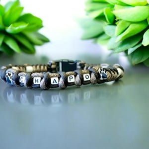 Può includere: Un braccialetto in paracord nero, grigio e beige con la parola "CHAPO" scritta con perline bianche. Il braccialetto presenta una chiusura nera e perline a forma di zampa.