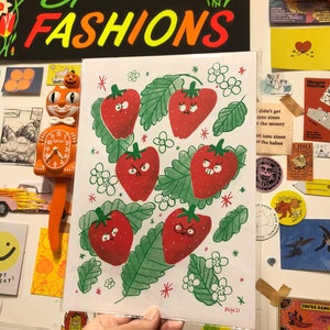 Puede incluir: Una tarjeta blanca con una ilustración verde, roja y blanca de fresas con caras enojadas. La tarjeta tiene un fondo blanco con estrellas rojas y blancas y flores blancas. El texto "ECHK21" está en la esquina inferior derecha de la tarjeta.