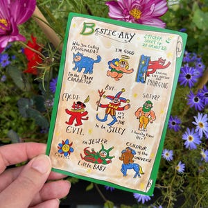 Puede incluir: Una hoja de pegatinas verde titulada "Bestie-ary" con ilustraciones de personajes de dibujos animados y frases como "I'm the Main Character" y "Living Life Outside the Margins". La hoja de pegatinas se sostiene contra un fondo de flores.