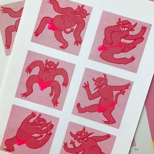 May include: A print featuring six red cartoon devils in various poses, each within a pink square. The devils have horns, tails, and are wearing pink briefs. The background is white, with additional prints visible.