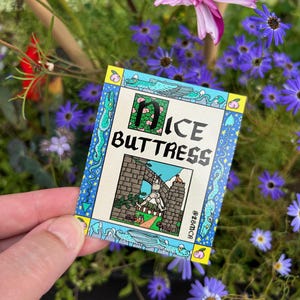 May include: A small rectangular card with the words "Dice Buttress" in a decorative font. The card features a colourful illustration of a medieval scene with a castle and a dragon. The border is blue with decorative elements.