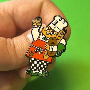 May include: An enamel pin featuring a cartoon chef with a white chef's hat, red apron, and checkered trousers. The chef is holding a baguette and a small round pastry. The pin is black and white with red, green, and brown accents.
