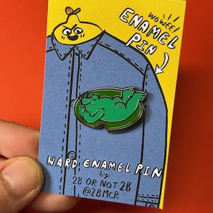 May include: A green enamel pin featuring a cartoon frog with a green body and black eyes. The pin is on a yellow and blue card with the text "WOWEE! ENAMEL PIN" and "HARD ENAMEL PIN by 2B OR NOT 2B @2BMCR".