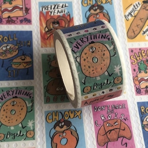 May include: A roll of washi tape with a colourful design featuring a cartoon bagel with the text "Everything Bagel" and "A  pple of my" on the tape. The tape is on a background of colourful patterned paper with images of various baked goods.