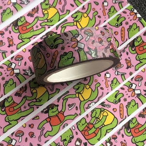Puede incluir: Un rollo de cinta adhesiva decorativa con un patrón repetido de ranas de dibujos animados, setas y pan sobre un fondo rosa. La cinta es marrón con un núcleo blanco. Las ranas están en varias poses.