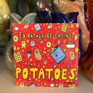 Pode incluir: Um cartão quadrado vermelho com o texto "I'd rather be eating potatoes" e várias ilustrações com tema de batata. O cartão apresenta batatas de desenho animado, batatas fritas e um saco de batatas fritas.