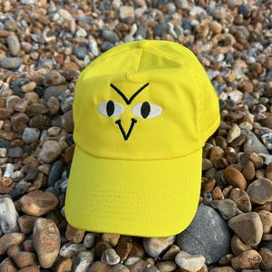 Könnte beinhalten: Eine gelbe Baseballkappe mit einem schwarzen gestickten Gesicht auf der Vorderseite. Das Gesicht hat zwei große weiße Augen, eine schwarze Stirnrunzel und einen schwarzen V-förmigen Mund.