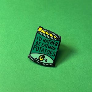 May include: Green enamel pin featuring a cartoon drawing of a bag of crisps. The bag reads "I'd rather be eating potatoes".