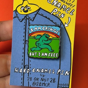 May include: A hard enamel pin featuring a green frog running with a rainbow and the text "I am cringe but I am free". The pin is on a blue background with the text "Hard Enamel Pin by 2B or Not 2B @2BMCR".