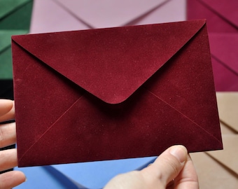 May include: A burgundy envelope is held in front of a collection of colorful envelopes. The envelope is made of a textured material and has a classic triangular flap. Other envelopes in shades of green, pink, purple, and tan are visible in the background.