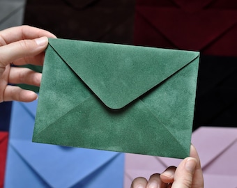 May include: A person holding a dark green envelope with a textured surface. The envelope is a muted shade of green and has a triangular flap. Other colorful envelopes are in the background.