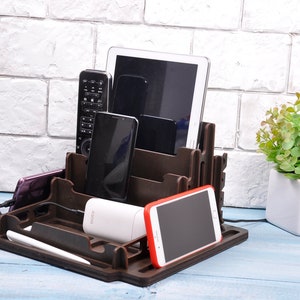 Op de afbeelding: Een bruine houten organizer met meerdere vakken om elektronische apparaten in op te bergen. De organizer heeft een tablet, telefoon en afstandsbediening erin. Er is een witte powerbank en een rode telefoon in de organizer.