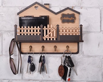 May include: Wooden key holder shaped like a house with a fence and a sign that says "Addington". It has five hooks for keys and a small shelf for mail or other items.