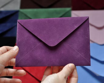May include: A deep purple envelope is held in front of a backdrop of various colored envelopes. The envelope is made of a textured paper and has a classic triangular flap. The background envelopes are in shades of blue, green, pink, and red.