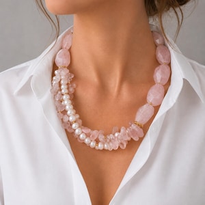 Puede incluir: Un collar llamativo de varias hebras con grandes pepitas de cuarzo rosa pálido facetadas y perlas blancas lisas más pequeñas. El diseño incorpora espaciadores de tono dorado entre las cuentas, creando un aspecto en capas alrededor del cuello.