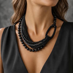May include: A multi-strand necklace featuring a combination of smooth black spherical beads and fine black seed bead strands. The design includes a prominent oval-shaped loop element on one side, connecting the various layers of beads into a cohesive piece.