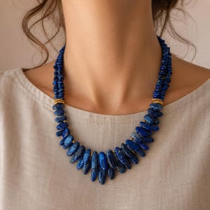 May include: A multi-strand necklace featuring deep blue lapis lazuli stones in elongated, irregular shapes. The stones are arranged in a cascading pattern, accented by small gold-toned spacers and two textured gold-toned cylindrical connectors.