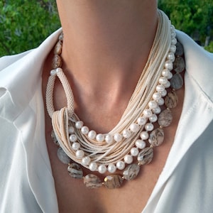 Puede incluir: Un collar de varias hebras con una variedad de cuentas y piedras. El collar incluye hebras de pequeñas cuentas blancas, perlas cremosas y piedras grandes, ovaladas y estampadas en tonos marrones y grises. El collar se usa sobre una camisa blanca.