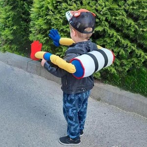 May include: A child wearing a black baseball cap and a black jacket, with a plush toy attached to their back and arms. The toy is primarily white with black stripes, red and blue accents, and yellow arms and hands.