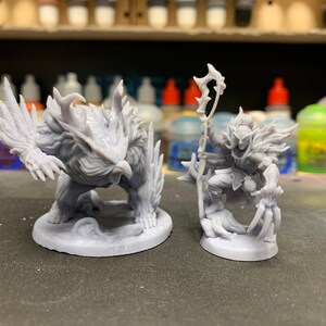 May include: Two gray 3D printed miniature figurines. One is a bird-like creature with sharp claws and feathers. The other is a humanoid figure with a staff and a cloak. Both figures are standing on circular bases.